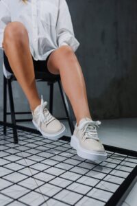 A person wearing a white long-sleeve shirt and black shorts sits on a black chair, showing off stylish shoes with thick soles. Their legs are crossed, giving the illusion that their feet look smaller against the dark, minimalist setting.