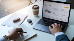 A person uses a laptop displaying car rental reviews, with a notepad, pen, coffee cup, car keys, and documents on the table. One hand types on the laptop while the other writes notes, suggesting research into essential tips for renting a car.